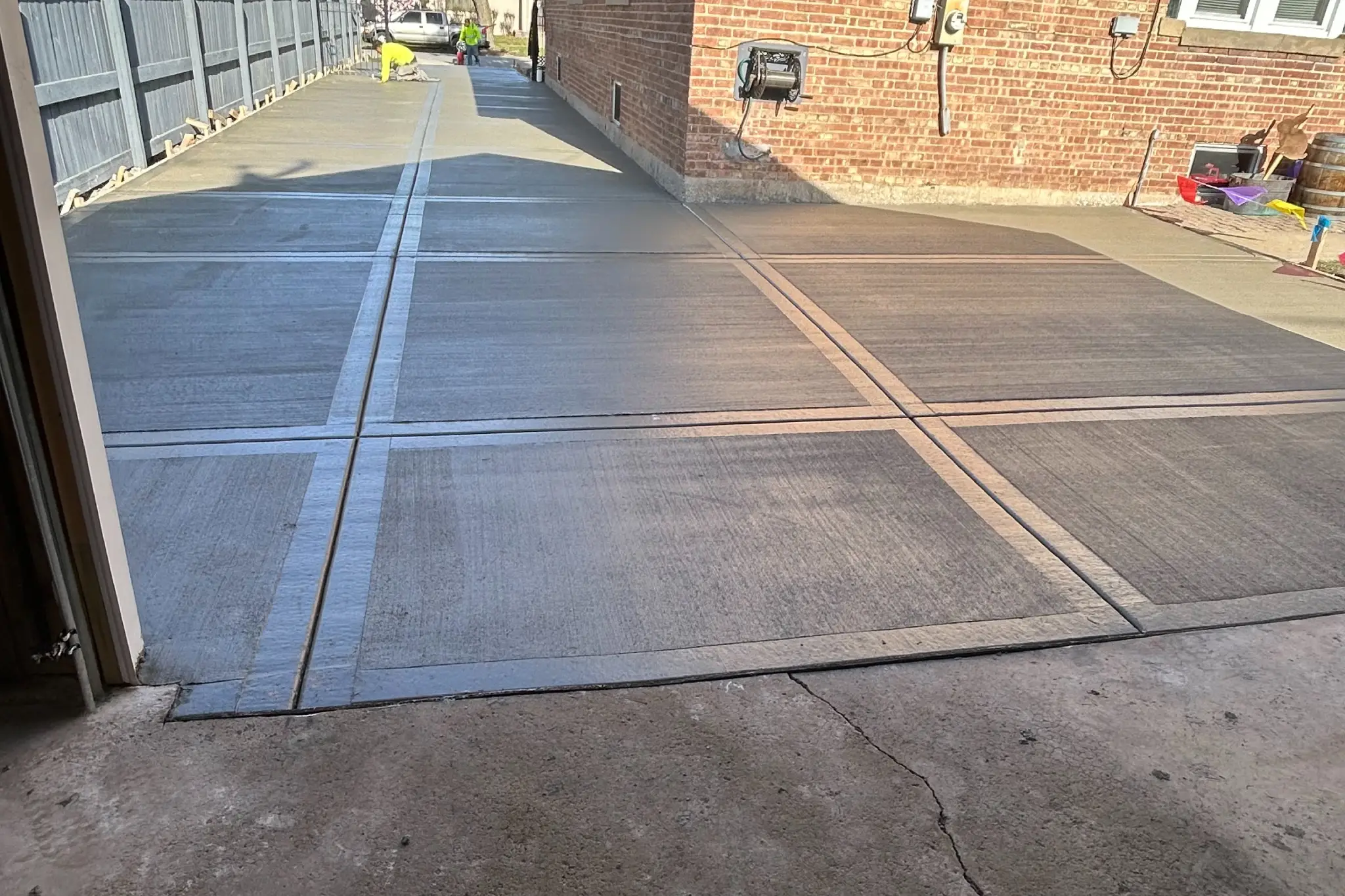 A view from a garage showing freshly poured concrete driveway, flanked by a red brick house and fence, under a clear blue sky