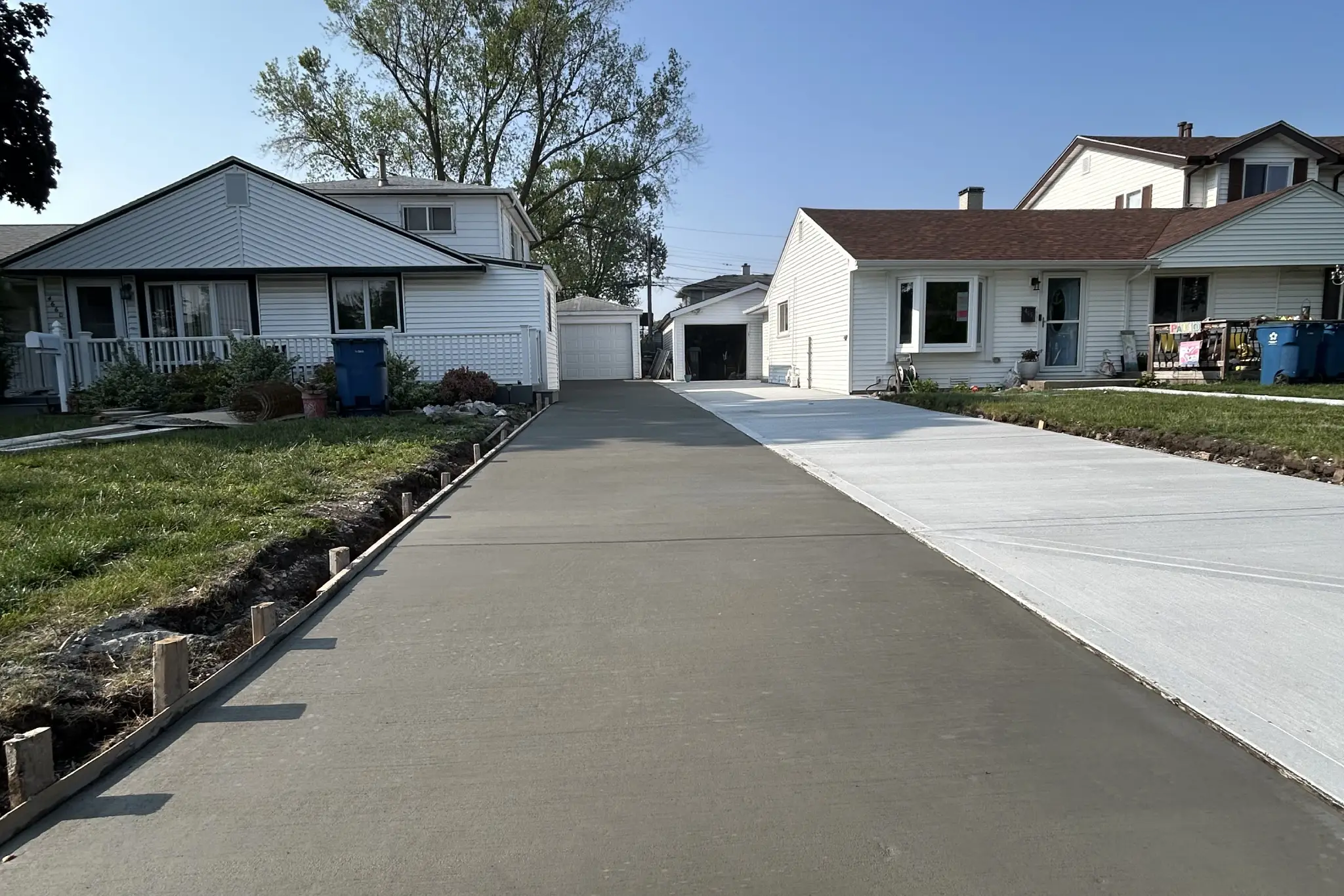 Freshly poured concrete driveway flanked by two houses under a clear blue sky, with wooden forms outlining the driveway edges