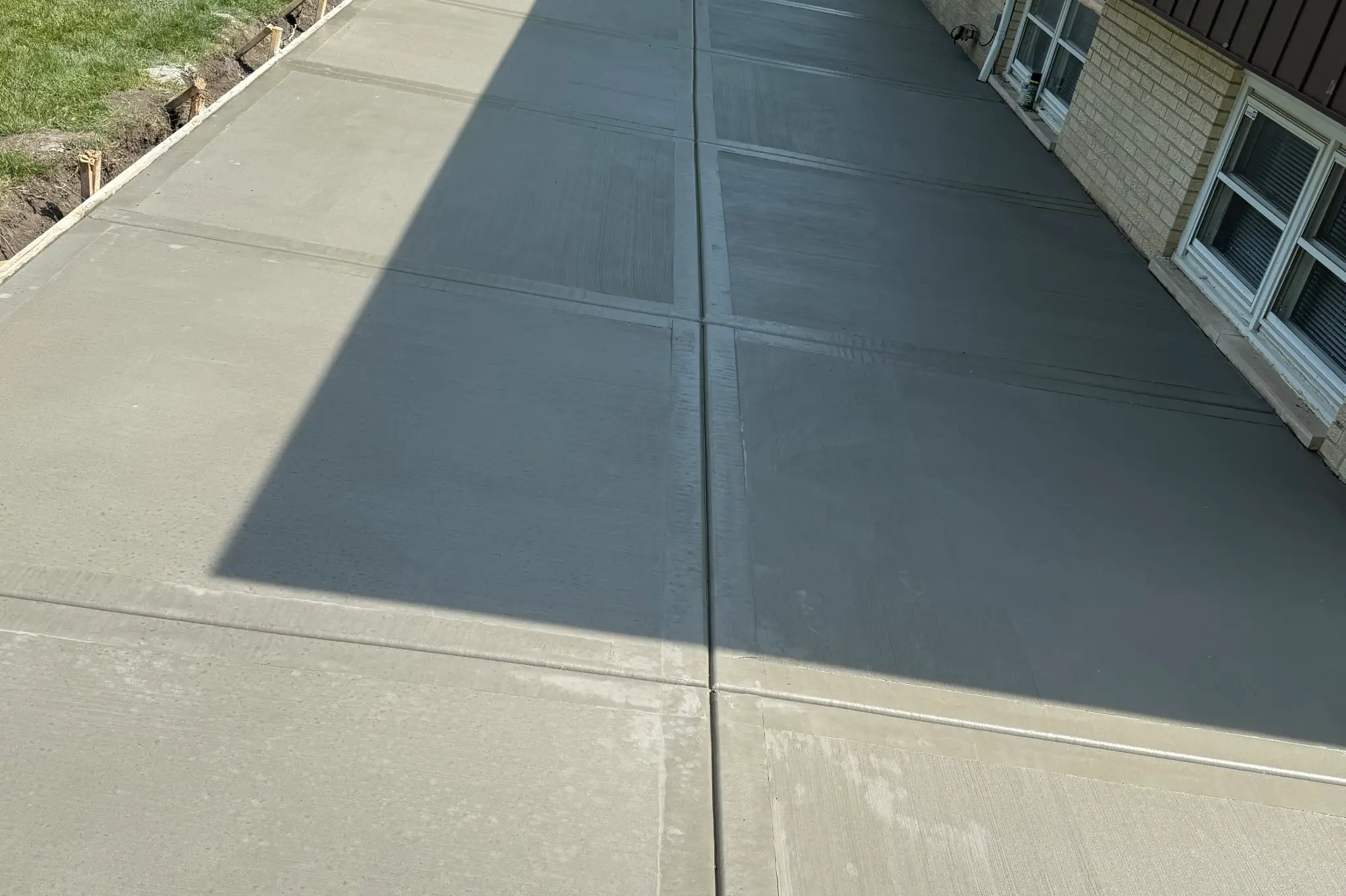 Freshly poured concrete patio with smooth surfaces, bordered by wooden forms and greenery, beside a house