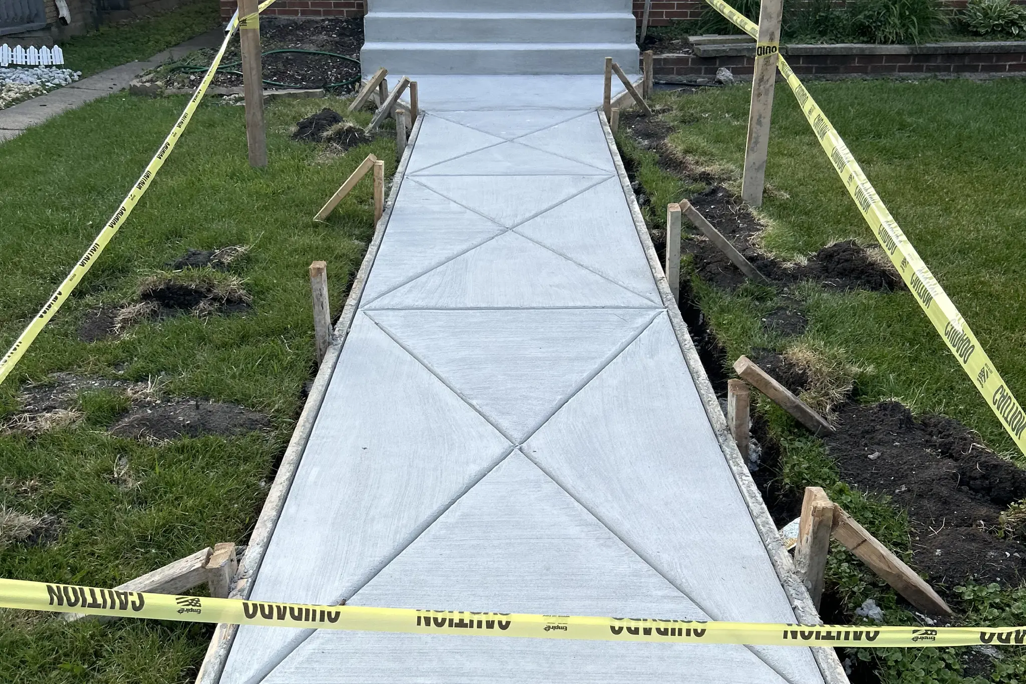 Freshly poured concrete sidewalk leading to a front door, bordered by caution tape and wooden supports, with grass patches on the sides