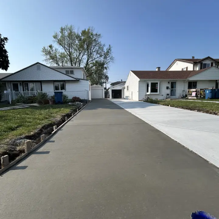 A newly poured concrete driveway stretches between two houses under a clear blue sky, framed by green grass and a tree