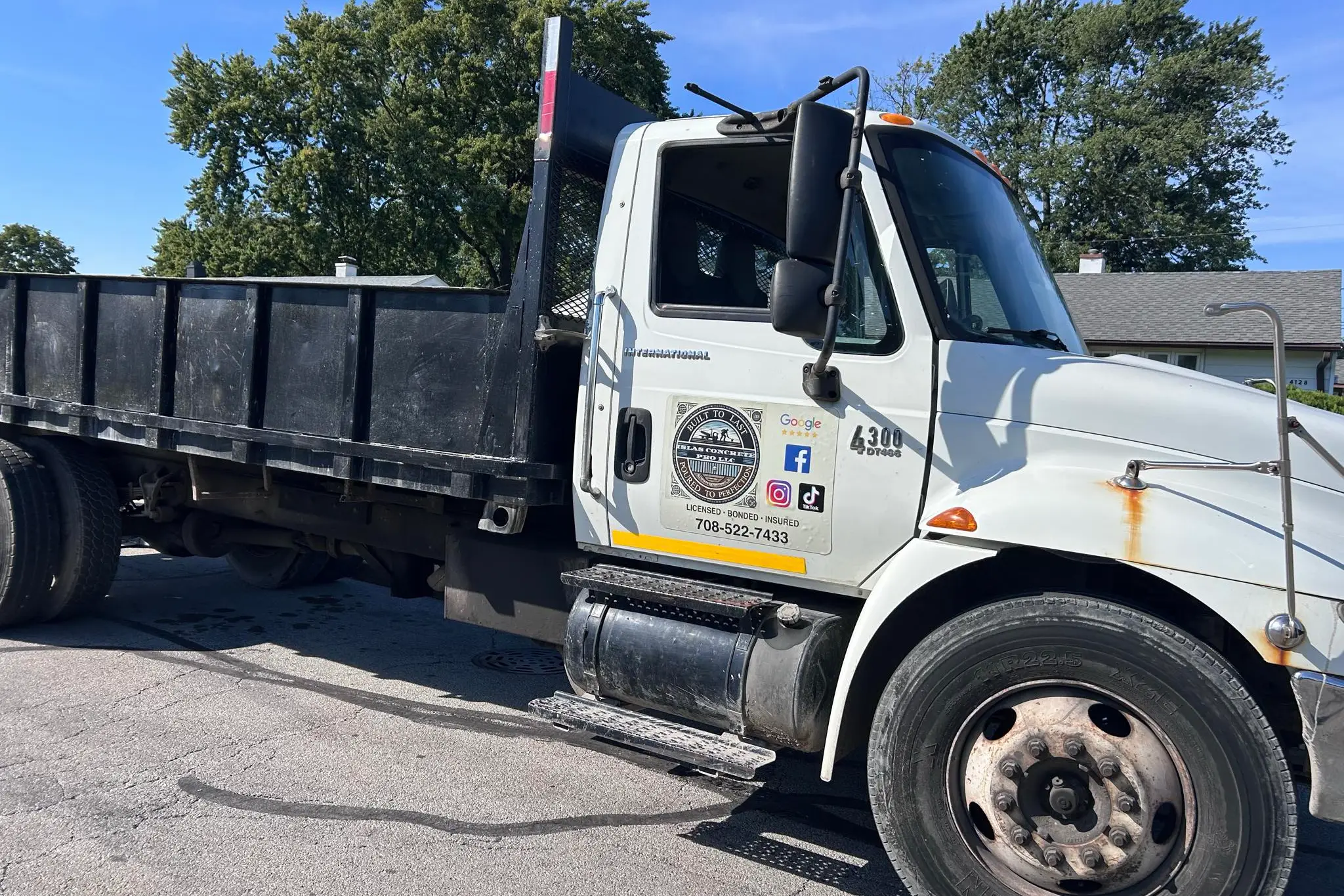 A white dump truck parked on the street, featuring a company logo and contact details on the side, under a clear blue sky