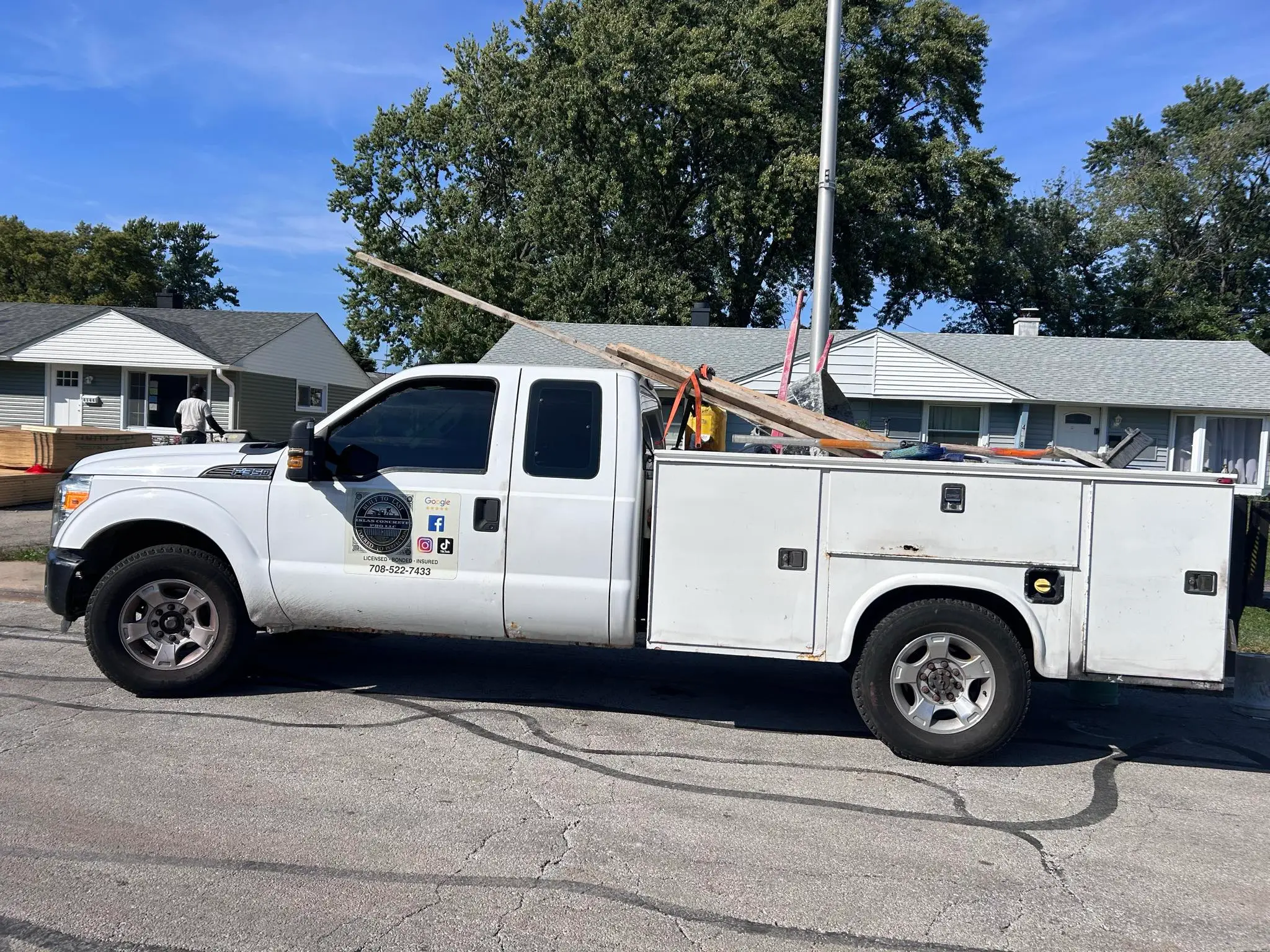 A white service truck parked on the street loaded with construction materials, with a house and trees in the background
