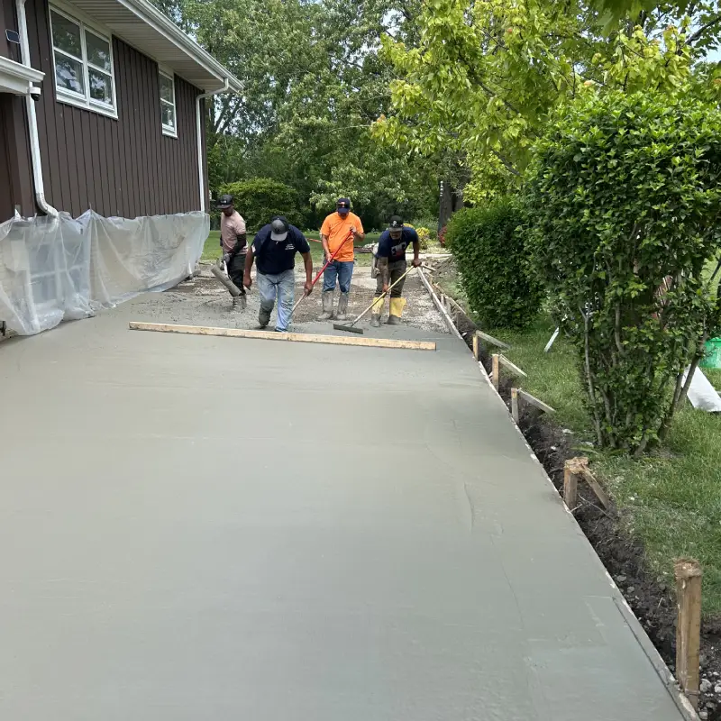 Four workers are pouring and smoothing wet concrete to create a new sidewalk beside a brown house under a clear blue sky